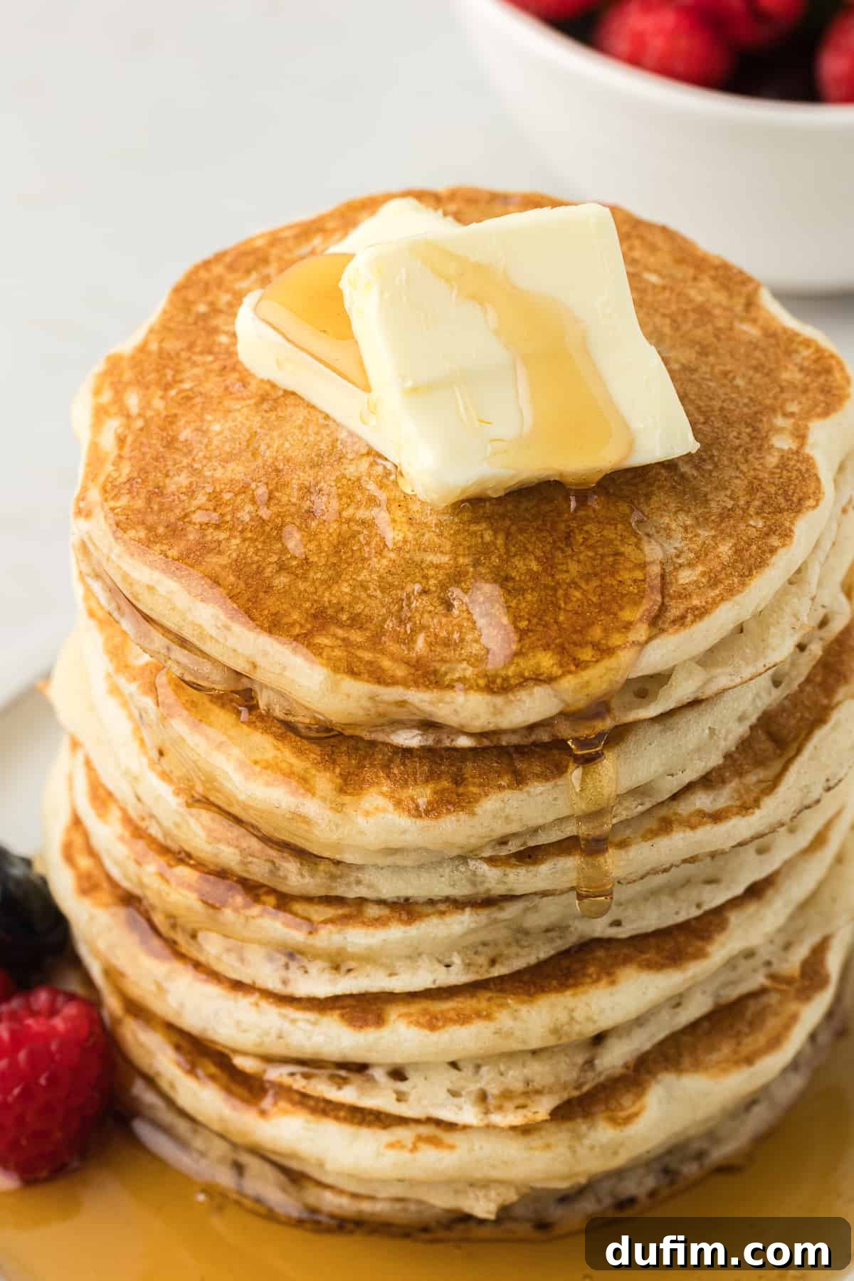 Simple Secrets to Fluffy Buttermilk Pancakes 13 A meticulously arranged white plate showcasing a tall, inviting stack of golden buttermilk pancakes, beautifully crowned with two creamy pats of butter. The plate is garnished with a vibrant array of fresh blueberries and raspberries, all glistening under a generous drizzle of rich maple syrup, creating a mouthwatering breakfast display.