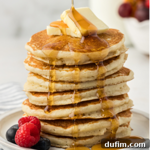 A tall stack of buttermilk pancakes on a white plate with two slices of butter on top and fresh blueberries and raspberries on the plate being drizzled with maple syrup