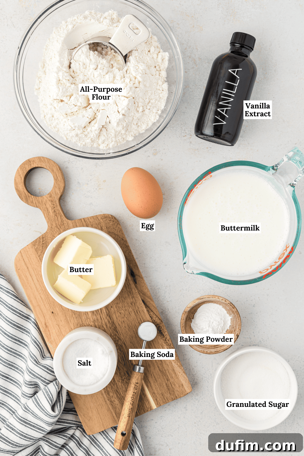 Simple Secrets to Fluffy Buttermilk Pancakes 3 An overhead view of carefully arranged ingredients for homemade buttermilk pancakes, including a bag of all-purpose flour, a bowl of granulated sugar, a bottle of vanilla extract, a carton of buttermilk, a single large egg, a stick of butter, and small measuring spoons for baking powder, baking soda, and salt, all ready for mixing.