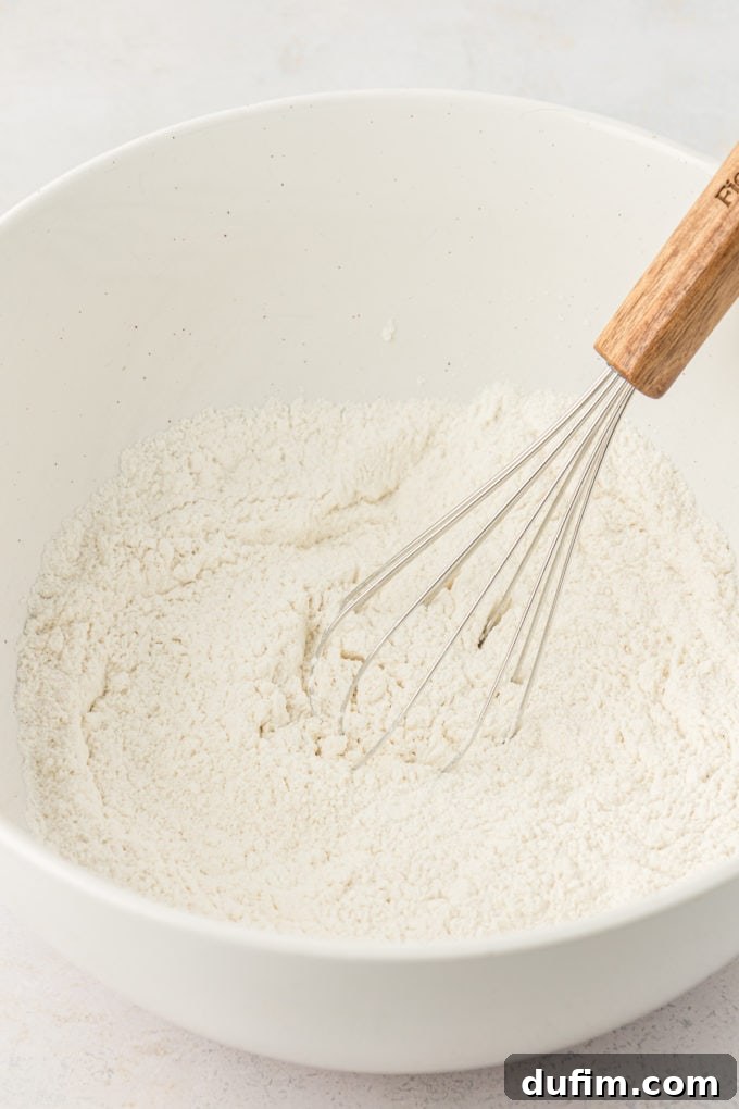 Simple Secrets to Fluffy Buttermilk Pancakes 5 Dry ingredients for buttermilk pancakes, including flour, sugar, baking powder, baking soda, and salt, neatly arranged in a white mixing bowl with a wire whisk poised inside, ready to be combined.