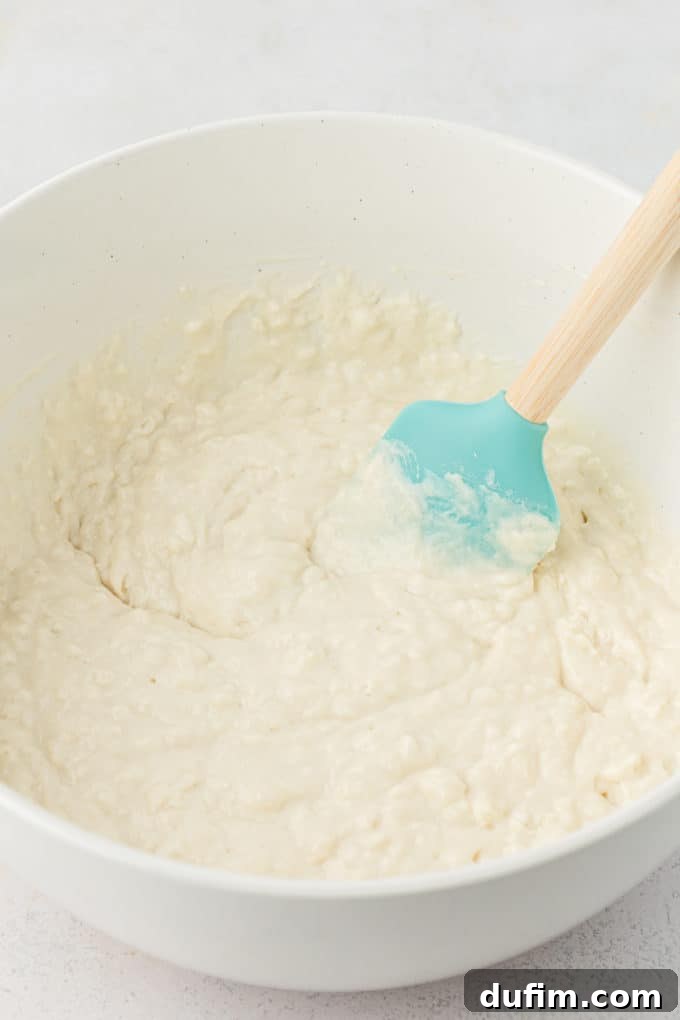 Simple Secrets to Fluffy Buttermilk Pancakes 6 Fluffy buttermilk pancake batter, with small lumps visible, being gently stirred in a white mixing bowl with a vibrant teal silicone spatula, illustrating the 'just combined' consistency.