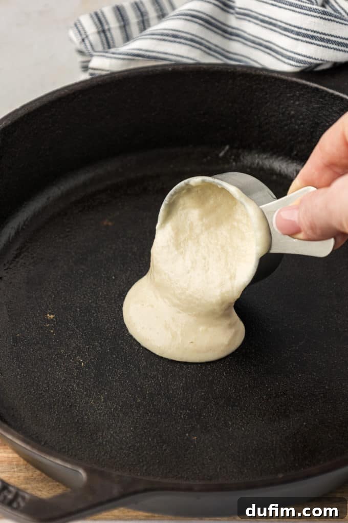 Simple Secrets to Fluffy Buttermilk Pancakes 7 Buttermilk pancake batter gracefully pouring from a measuring cup onto a sizzling cast iron skillet, creating a perfectly round pancake, with a blue and white striped kitchen towel draped nearby.