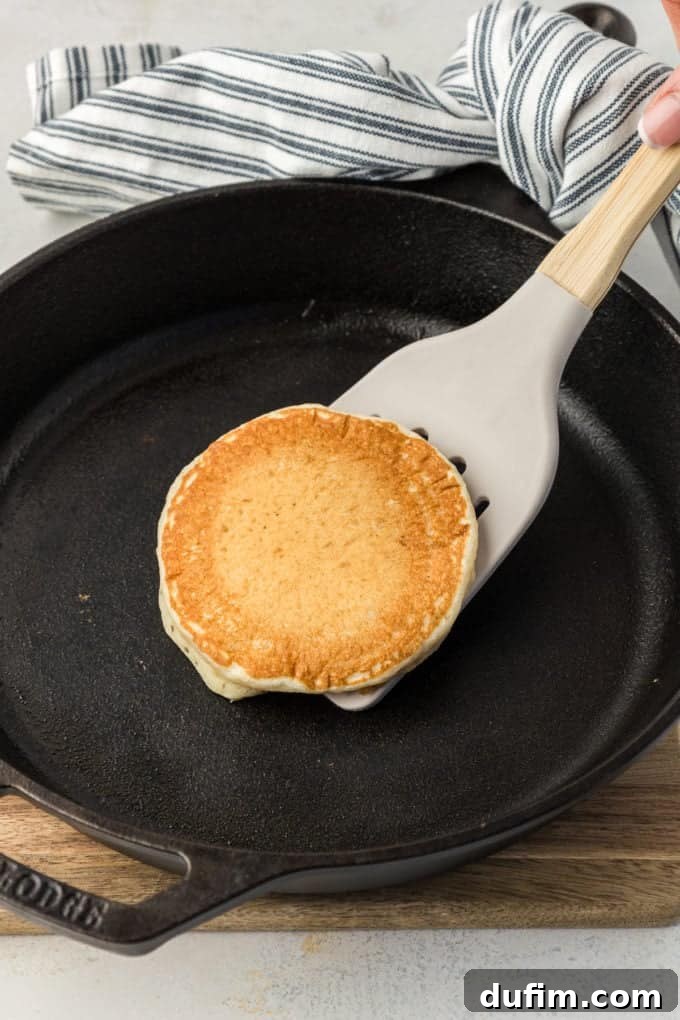 Simple Secrets to Fluffy Buttermilk Pancakes 8 A golden-brown buttermilk pancake being expertly flipped mid-air with a spatula over a hot cast iron skillet, with a blue and white striped towel gripping the handle, showcasing the perfect moment for turning.