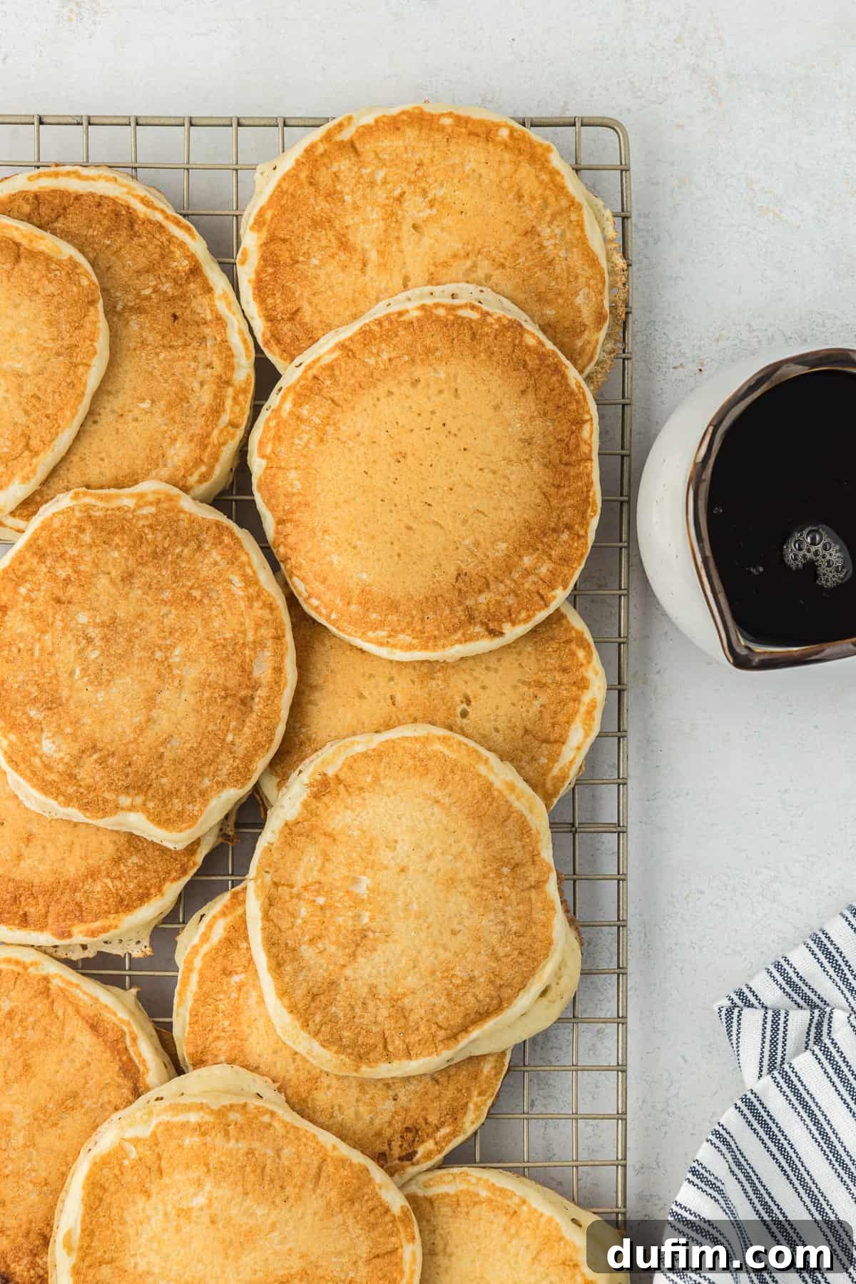 Simple Secrets to Fluffy Buttermilk Pancakes 9 A cooling rack filled with freshly cooked, golden-brown buttermilk pancakes, steaming gently, beside a small bowl of maple syrup and a blue and white striped kitchen towel, ready for serving.