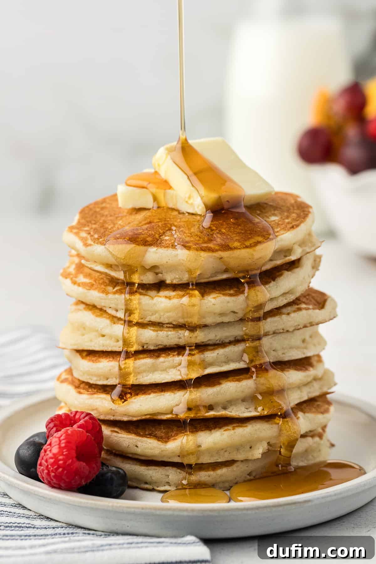 Simple Secrets to Fluffy Buttermilk Pancakes 10 A majestically tall stack of five fluffy buttermilk pancakes, adorned with two melting pats of butter, a generous scattering of fresh blueberries and raspberries, and cascades of rich maple syrup, all resting on a pristine white plate.