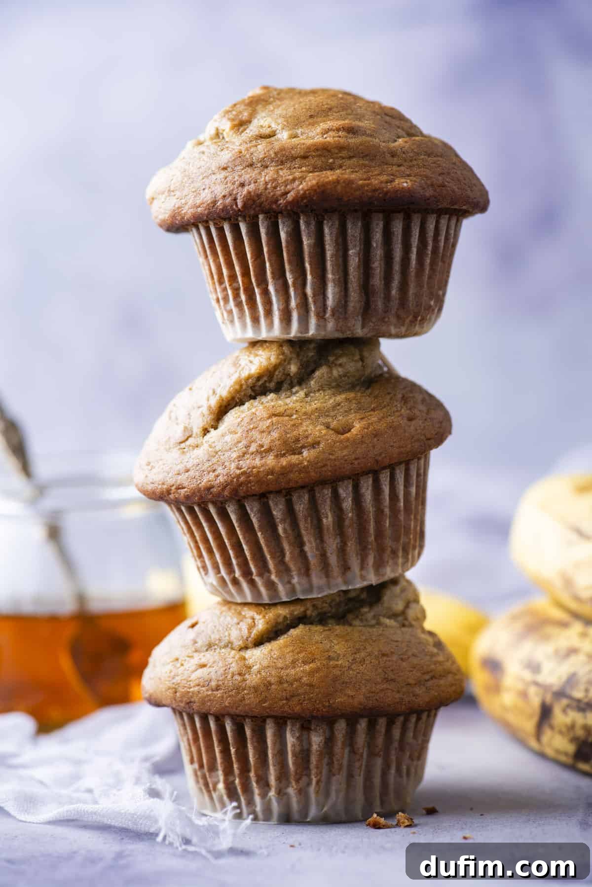 Three healthy banana muffins stacked in a tower on a vibrant purple background, showcasing their fluffy texture and perfect domes.