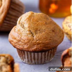 A close-up of a golden-brown healthy banana muffin in a paper wrapper, surrounded by other muffins on a light gray surface, with a blurred jar in the background.