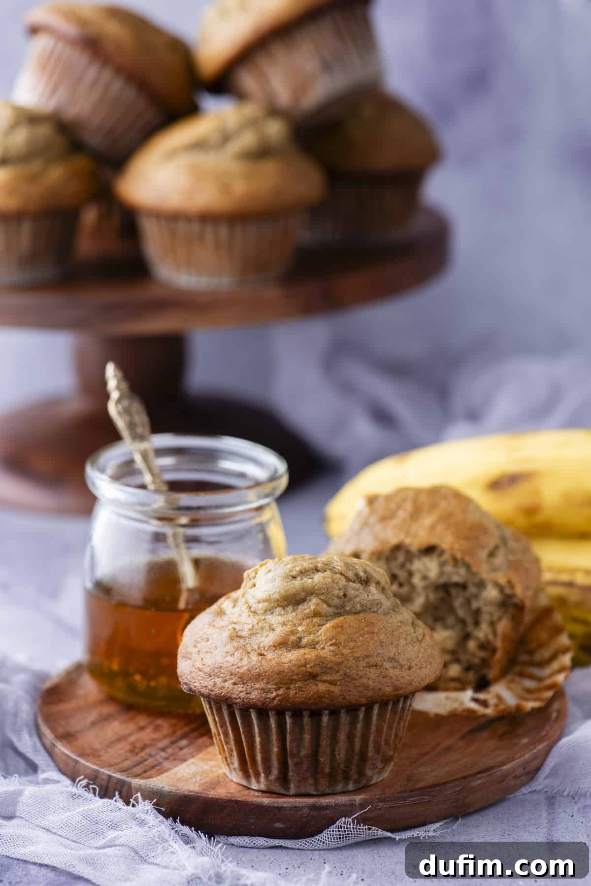 Healthy banana muffins displayed on a wooden plate, with a jar of honey and a muffin with a bite taken out, emphasizing its appealing texture and natural sweetness.