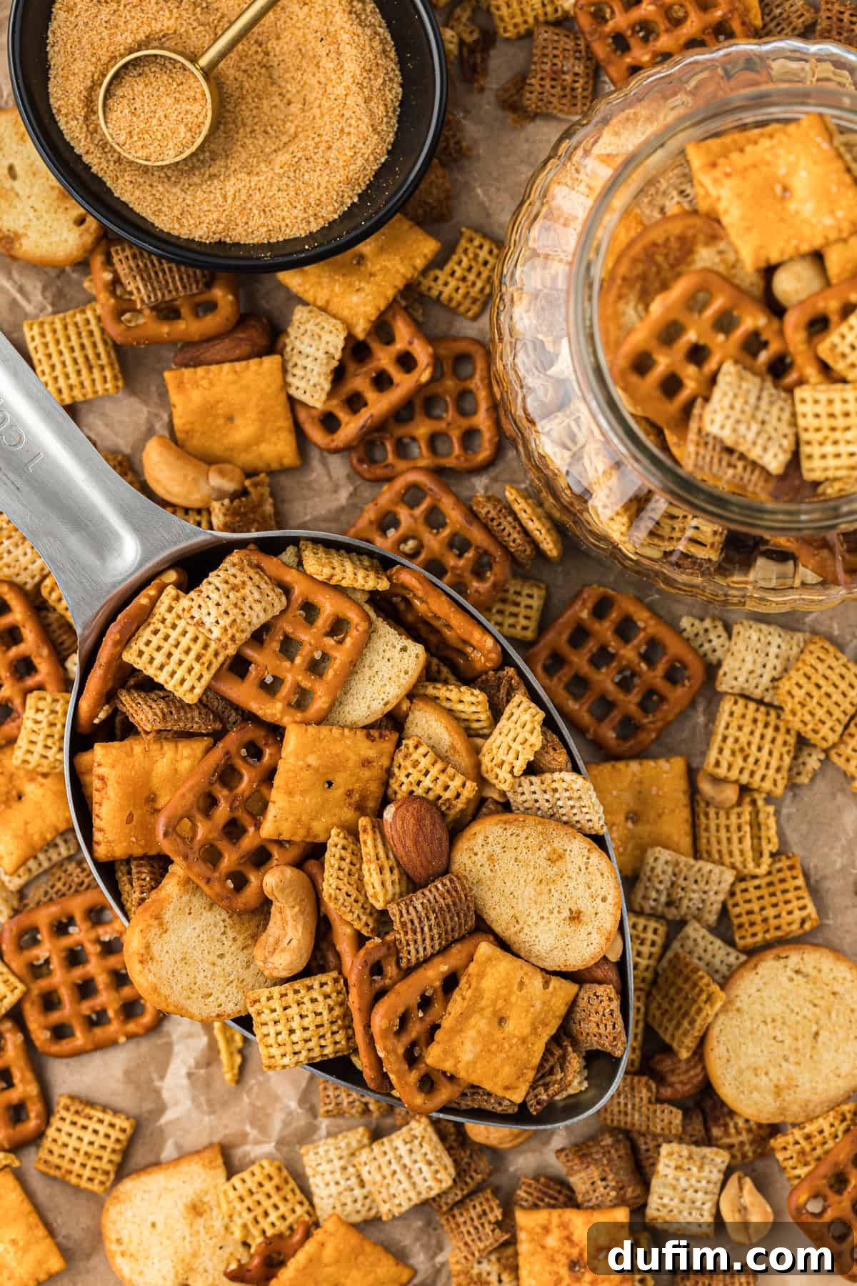 Unlock the Secret to Perfect Chex Mix 2 Overhead view of a large metal scoop full of golden-brown homemade Chex Mix, with more mix scattered on a rustic brown paper surface. A small black bowl of seasoning and a clear glass jar of Chex Mix are also visible.