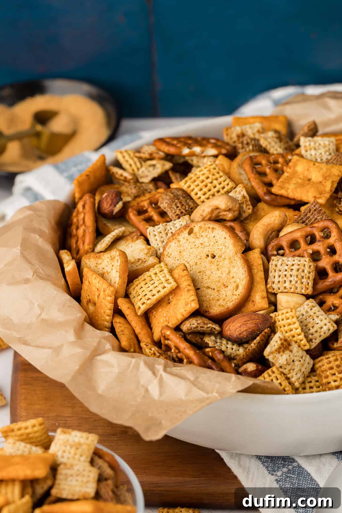 Unlock the Secret to Perfect Chex Mix 12 A white bowl lined with brown paper, brimming with homemade Chex Mix, rests on a wooden board next to a white and blue striped towel. A smaller bowl of Chex Mix and a bowl of seasoning are blurred in the background, creating a cozy and inviting scene.
