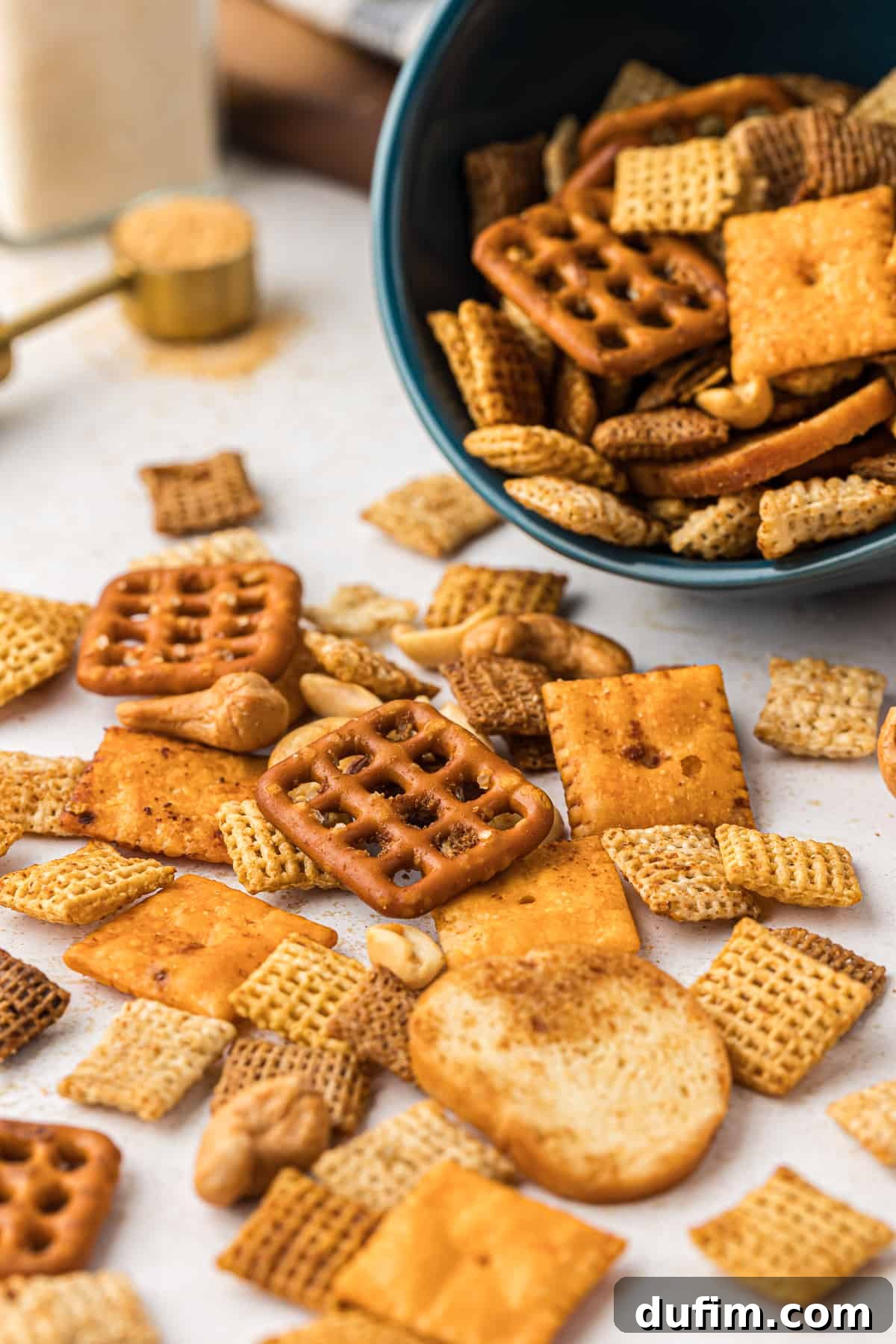 Unlock the Secret to Perfect Chex Mix 4 Homemade Chex Mix spread out on a clean white surface, with a dark teal bowl full of the snack mix tilted beside it, inviting a closer look at its perfectly seasoned pieces.