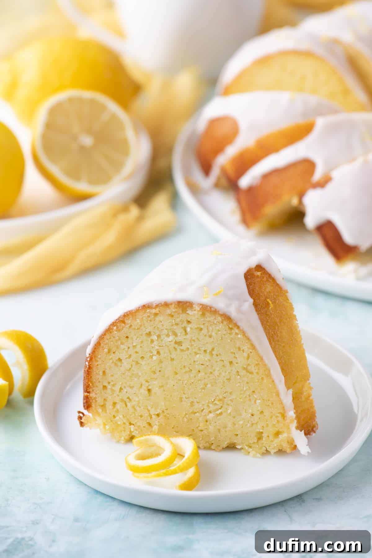 A beautifully sliced lemon bundt cake on a small, round white plate, garnished with fresh lemon peel, with more cake slices and fresh lemons artfully arranged in the background.