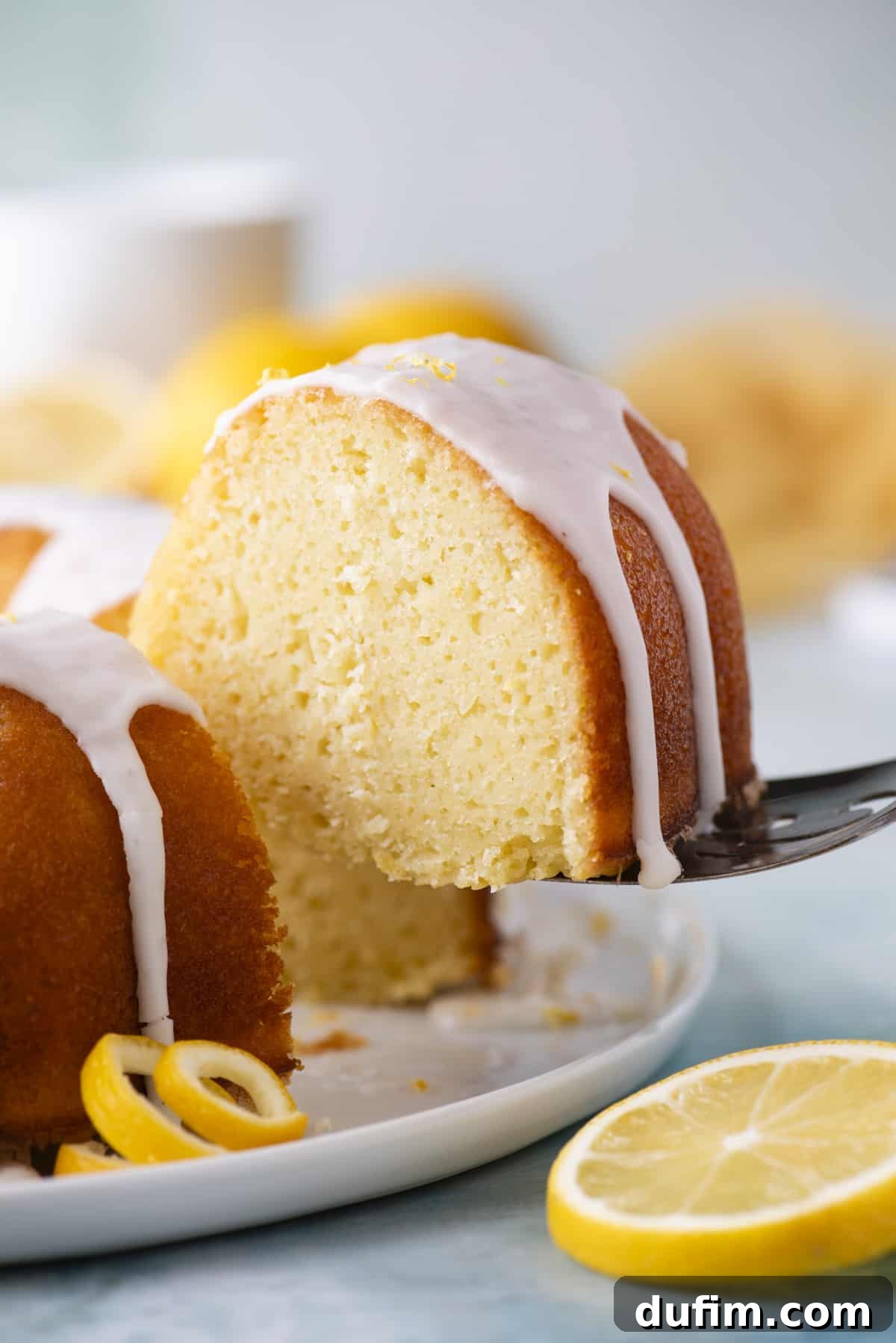 A tempting slice of lemon bundt cake being carefully lifted from the main cake on a pristine white plate, resting on a serene blue surface. A vibrant fresh lemon slice sits gracefully beside it, adding to the visual appeal.