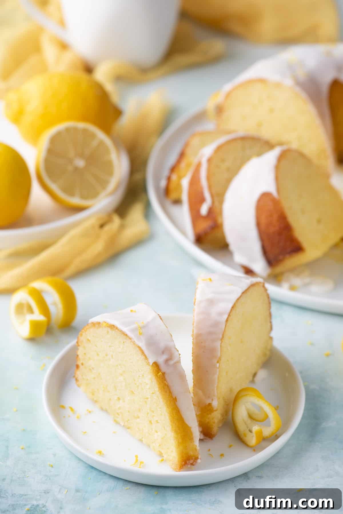Two inviting slices of lemon bundt cake arranged on a small white plate, surrounded by delicate fresh lemon peels. Nearby, another white plate holds additional cake slices, and a third displays vibrant fresh lemons, creating a delectable and appealing spread.