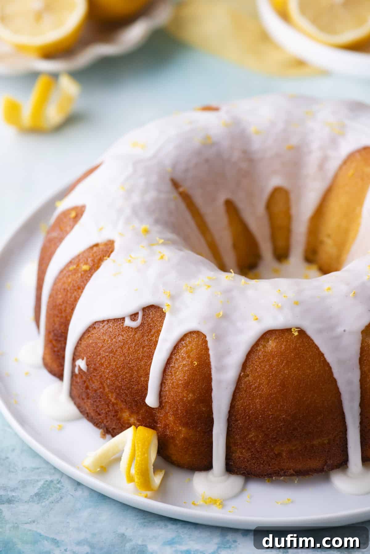 A magnificent lemon bundt cake, still on its round, white plate and garnished with fresh lemon peels, elegantly positioned on a blue surface. Small white plates holding fresh lemons, and more slices and peels, surround the cake in a vibrant display.
