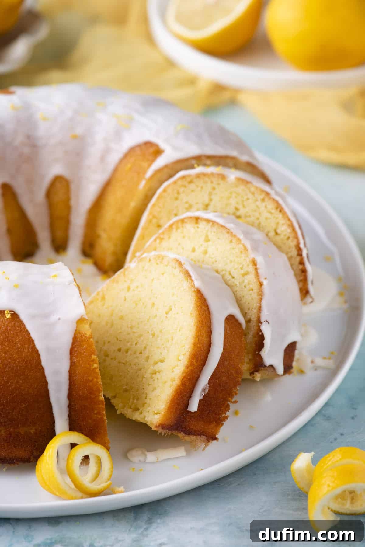 A beautifully presented lemon bundt cake on a round, white plate, with three slices already cut and the remaining cake whole. It is adorned with fresh lemon peels, and a small white plate nearby holds additional fresh lemons, creating an elegant and inviting dessert spread.