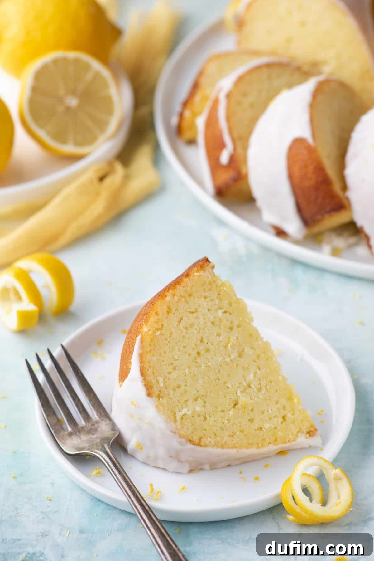 A perfectly portioned slice of lemon bundt cake resting on a small white plate, accompanied by a fork and surrounded by fresh lemon peels. In the background, two more white plates are visible, one holding additional cake slices and the other displaying whole fresh lemons, creating an inviting spread.