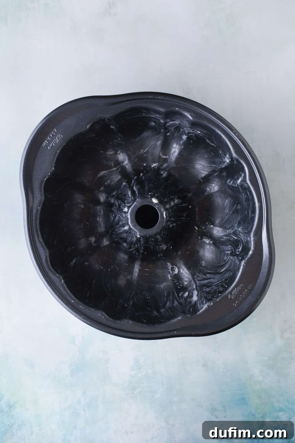 An overhead view of a thoroughly greased bundt cake pan, ready for batter, resting on a light blue surface.