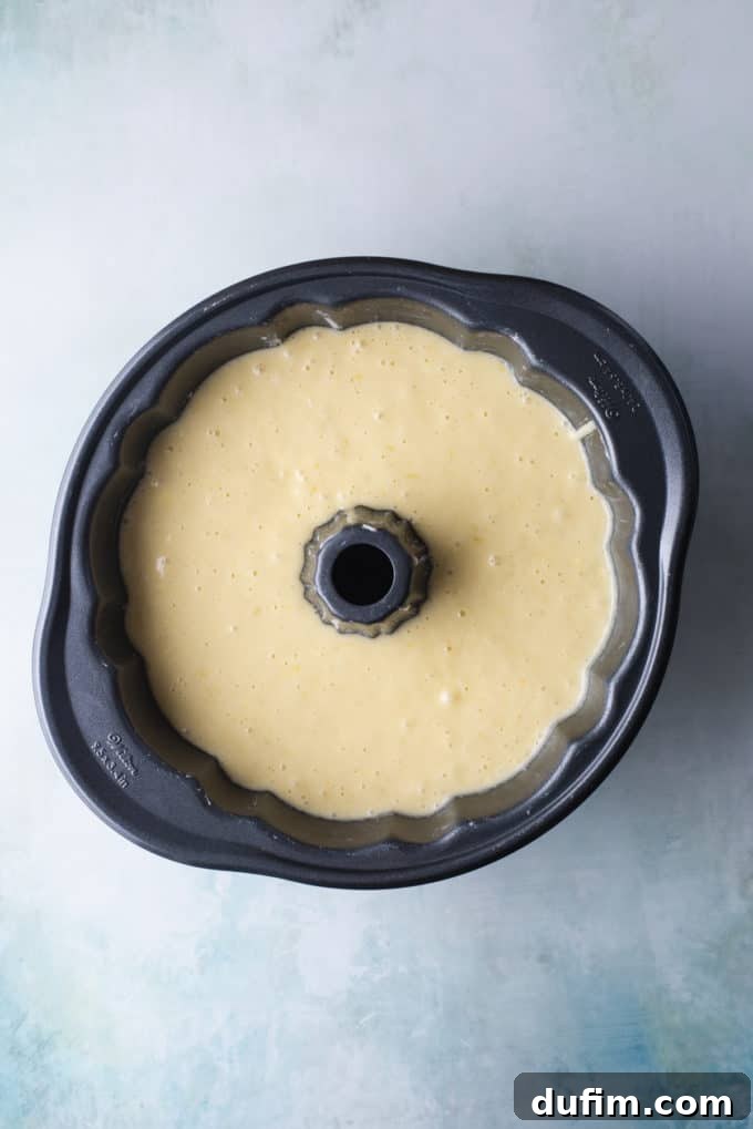 An overhead view of smooth lemon bundt cake batter neatly poured into a perfectly prepared bundt pan, sitting on a light blue surface, ready for baking.