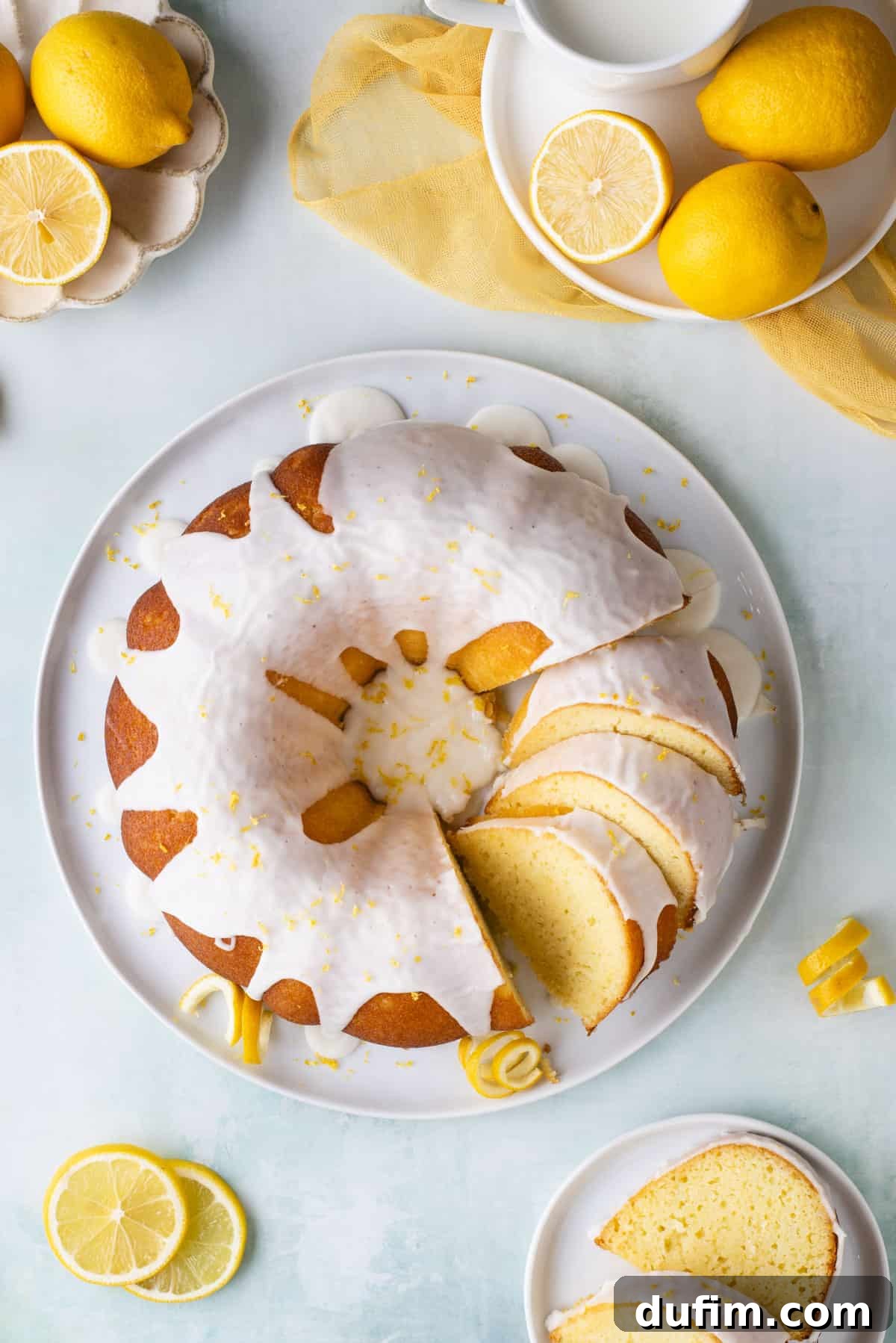 An inviting overhead view of a beautifully glazed lemon bundt cake resting on a round, white plate. Three perfect slices have been cut, while the rest of the cake remains whole, ready for serving. It is gracefully surrounded by smaller white plates, one holding two more slices of lemon bundt cake, and another showcasing vibrant fresh lemons.