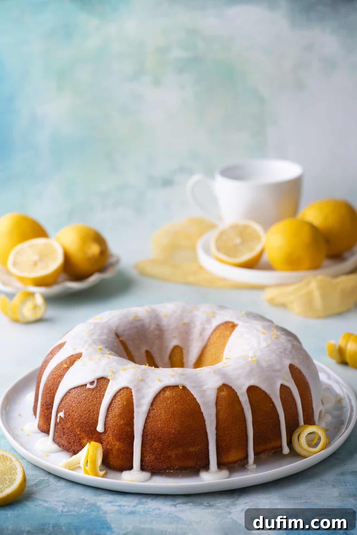A beautifully finished lemon bundt cake on a round, white plate, adorned with fresh lemon peels, all resting on a charming blue surface. Small white plates holding fresh lemons and more lemon slices and peels are arranged around it, completing a delightful display.