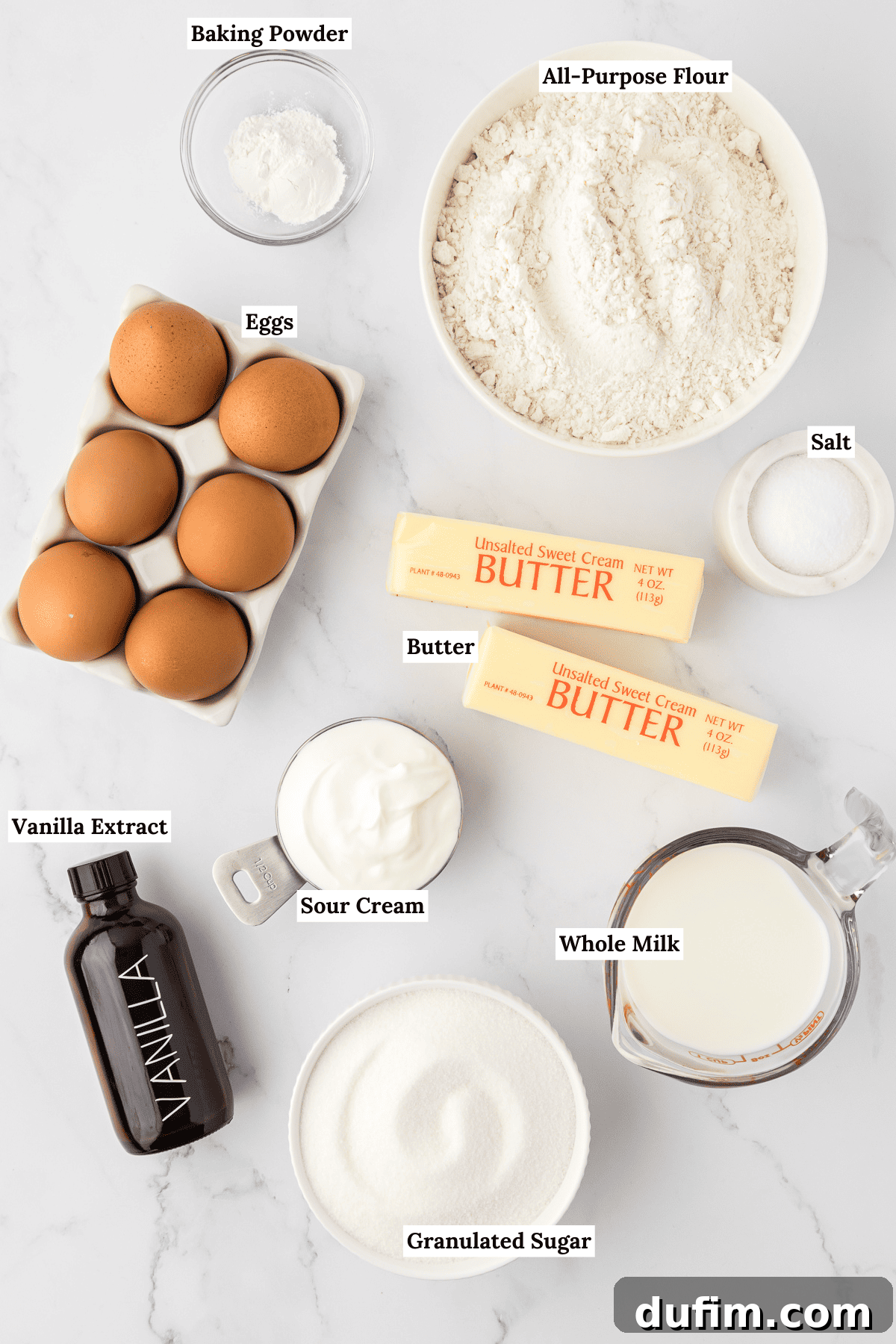 overhead view of the ingredients for bundt cake including a small glass bowl of baking powder, a white bowl of all-purpose flour, a carton of 6 eggs, a small white bowl of salt, two sticks of butter, a bottle of vanilla extract, a small measuring scoop of sour cream, a glass measuring cup of whole milk, and a white bowl of granulated sugar