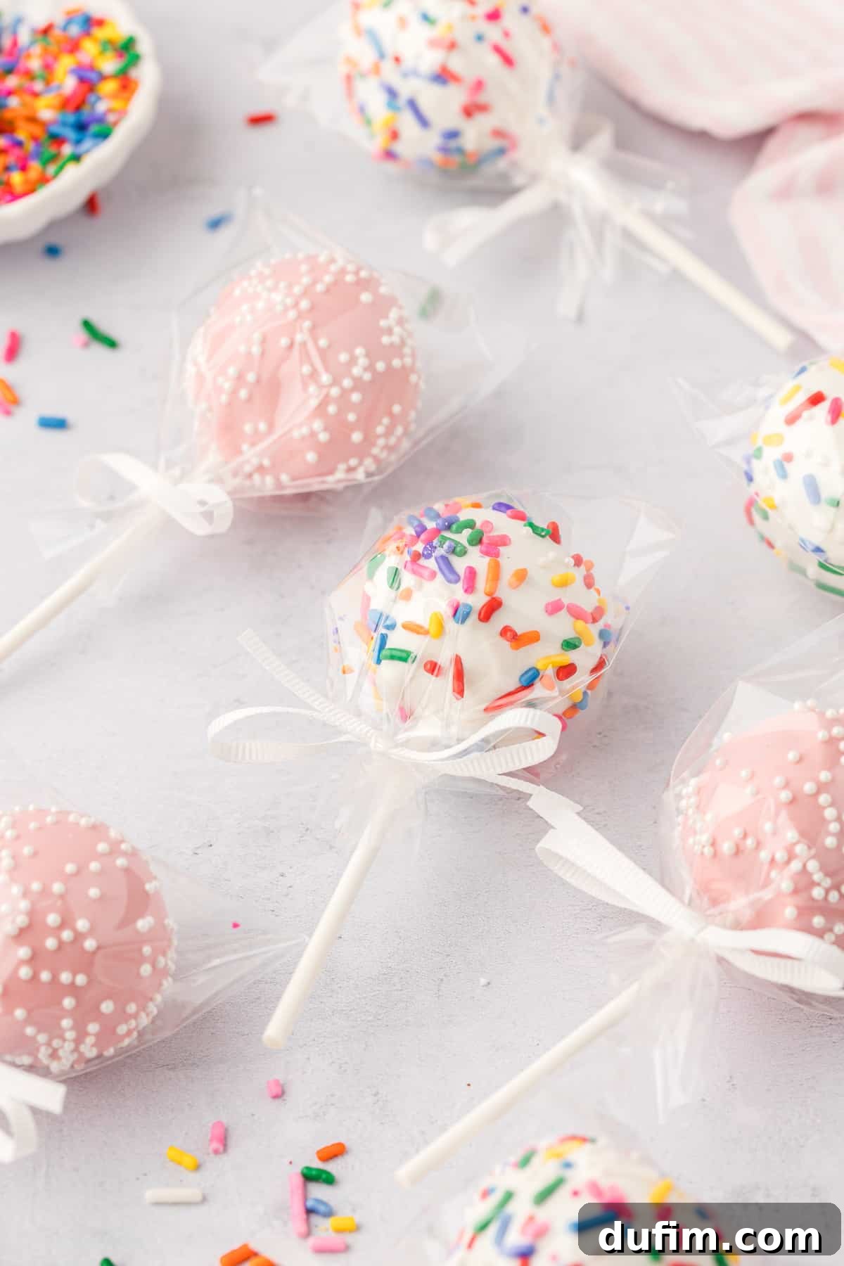 Individually wrapped homemade cake pops on a light grey surface. Some are vibrant pink with delicate white sprinkles, others are classic white with festive rainbow sprinkles. More rainbow sprinkles are scattered around, adding a playful touch. These perfectly crafted treats are ideal for gifting or party favors, ensuring a delightful surprise.