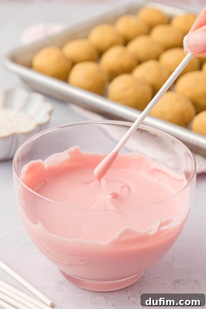 A lollipop stick, glistening with freshly dipped pink candy melts, is held poised directly over a clear glass bowl brimming with more smooth, melted pink coating. In the background, a baking sheet of plain, un-dipped cake pops patiently awaits its turn, illustrating the immediate next step in the cake pop creation process.