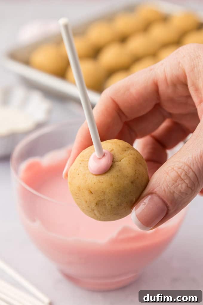 A careful hand firmly holds a cake pop on a stick, showcasing the delicate ring of melted pink candy melts that securely seals the stick to the cake ball. In the background, a bowl of more pink candy melts and a baking sheet of plain cake pops are visible, illustrating the preparation for the full dipping process.