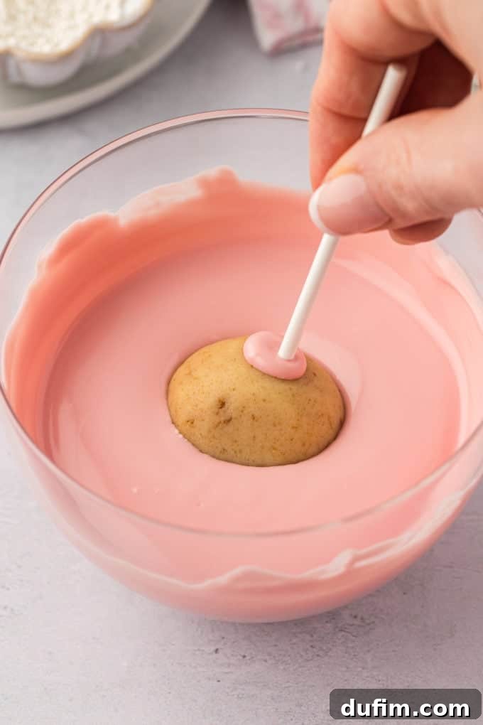 A pristine pink cake pop, affixed to a white stick, is being carefully and smoothly dipped into a clear glass bowl filled with vibrant, melted pink candy melts. The coating covers the cake pop flawlessly, creating a uniform and attractive surface.