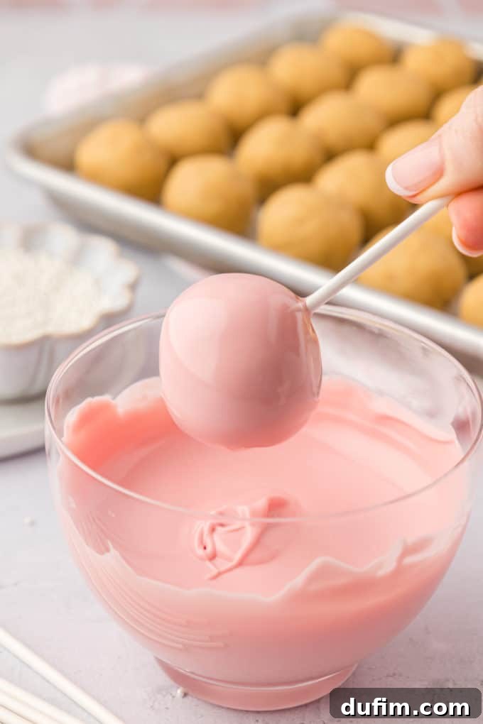A freshly dipped pink cake pop, exhibiting a flawless, shiny coating, is held momentarily over a clear glass bowl filled with more melted pink candy melts. In the background, a baking sheet of plain cake balls patiently waits. This image captures the pristine appearance of a perfectly coated cake pop, ready for its final decorative touches and setting.