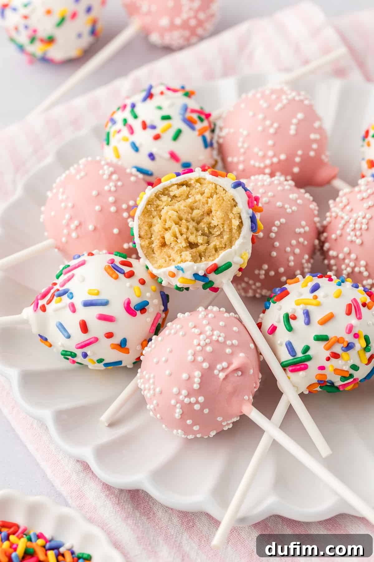 A charming white plate holds a delightful and inviting pile of assorted cake pops. Some are a soft, appealing light pink adorned with delicate white sprinkles, while others are a classic white embellished with festive rainbow sprinkles. The top cake pop, nestled invitingly, has a tempting bite taken out, proudly showcasing its incredibly moist and rich interior texture.