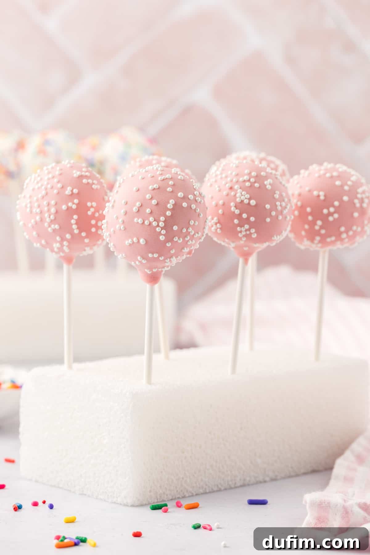 Six adorable pink cake pops, each adorned with delicate white sprinkles, stand perfectly upright in a clean white styrofoam block. A stylish white and pink striped towel is draped nearby, and scattered rainbow sprinkles add a charming, festive touch to the entire arrangement. This image exudes a delightful and celebratory atmosphere.