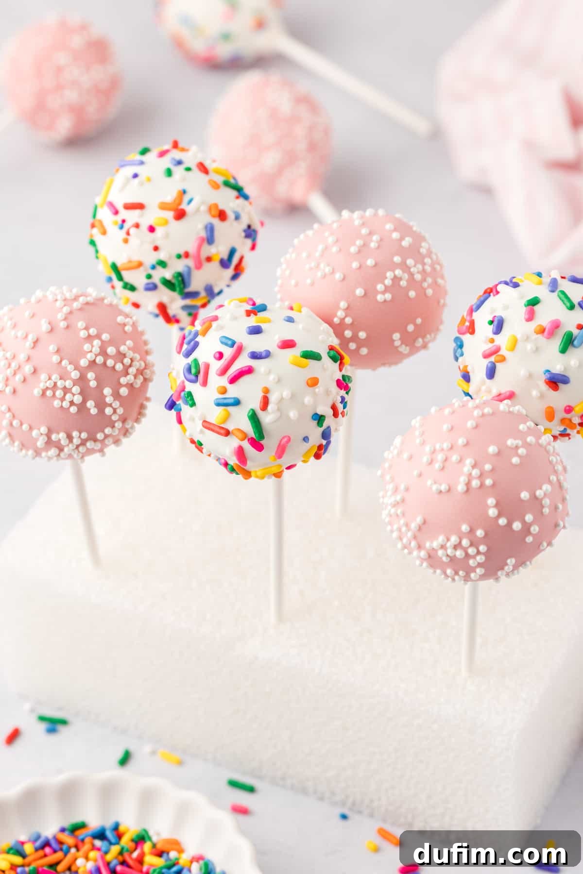 Six vibrant cake pops, three elegant pink with white sprinkles and three cheerful white with rainbow sprinkles, standing upright in a clean styrofoam block. A small white dish of rainbow sprinkles sits in the foreground, with more finished cake pops subtly blurred in the background, creating a festive and inviting scene.