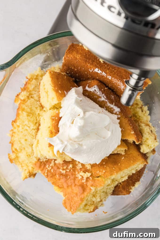 A generous, clean scoop of creamy white frosting delicately placed on top of the crumbled cake pieces, nestled within an electric mixing bowl. This visual signifies the crucial moment before blending, where cake and frosting unite to form the perfect cake pop dough.