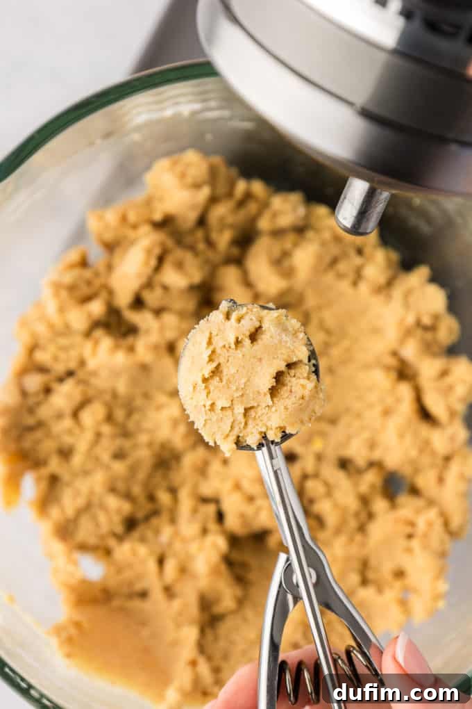 A tablespoon-sized cookie scoop, perfectly filled with the prepared cake pop mixture, is held gracefully over a mixing bowl full of more cake pop dough. This close-up illustrates the ideal consistency of the mixture, ready for shaping.