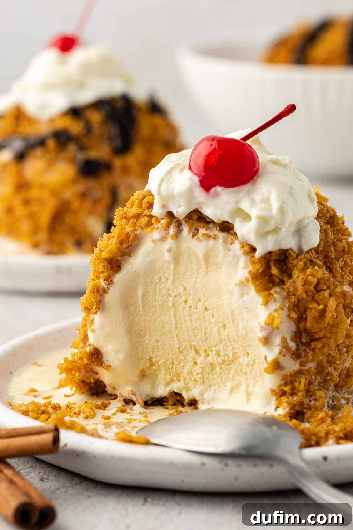 A perfectly prepared no-fry fried ice cream ball on a white plate, topped with a generous swirl of whipped cream and a bright red maraschino cherry. A spoon rests beside it, inviting a delicious bite. In the soft-focus background, another plate of ice cream and a bowl hint at more delectable treats.