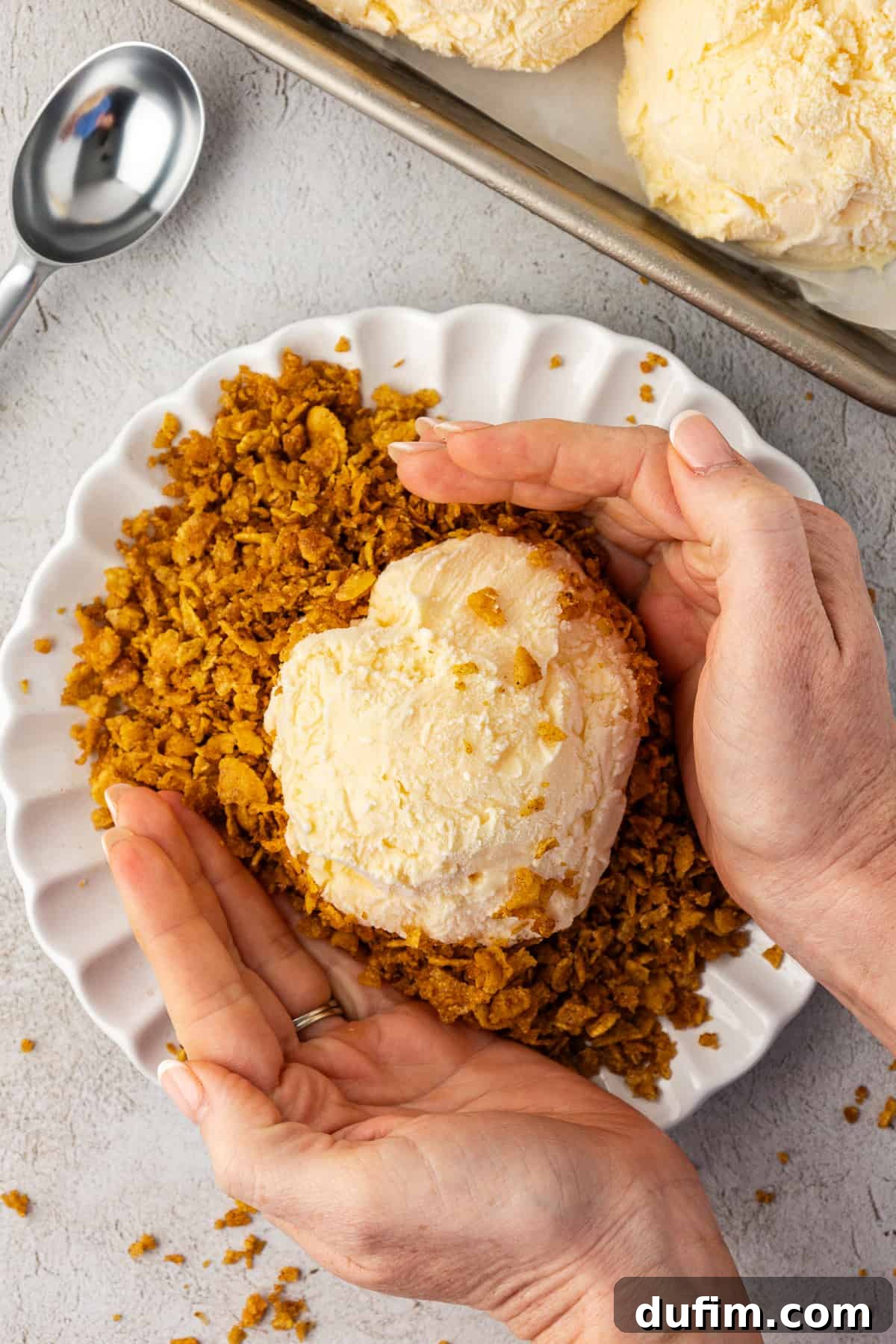 Two hands are carefully pressing the delicious cornflake cereal mixture onto a ball of vanilla ice cream, ensuring an even and complete coating. A sheet pan with more ice cream balls is visible in the background.