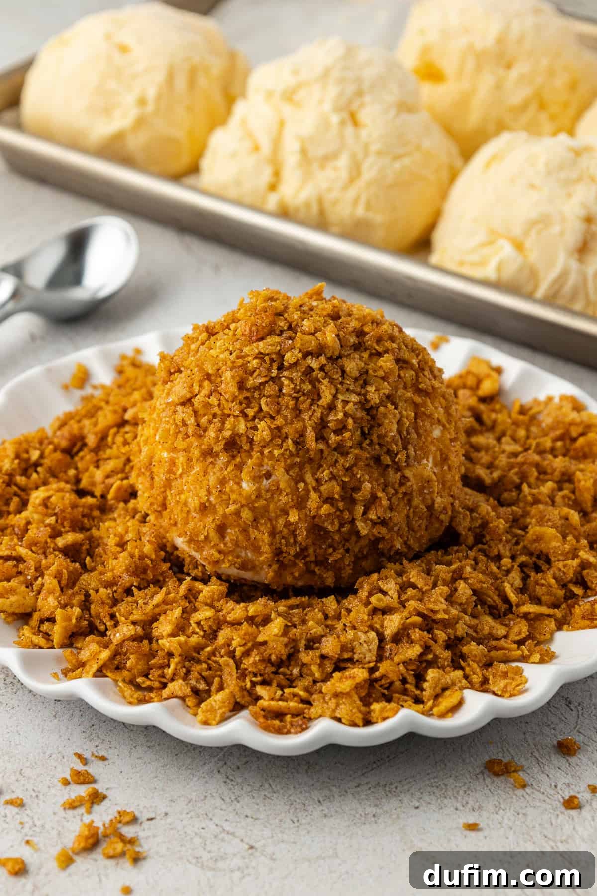 A perfectly coated ice cream ball, covered in the crunchy cornflake cereal mixture, rests on a bed of extra crumbs on a white plate, positioned next to a sheet pan displaying rows of plain vanilla ice cream balls ready for their coating.