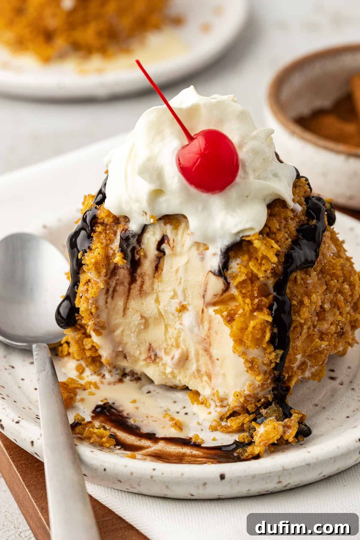 A close-up of a single fried ice cream ball on a small white plate with a spoon, generously topped with chocolate syrup, whipped cream, and a bright red cherry. A bite has been taken, revealing the creamy interior.