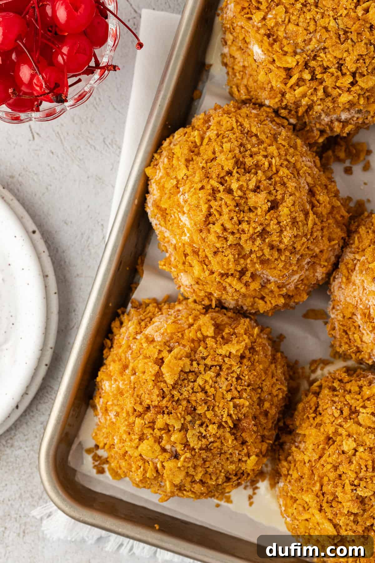 Multiple no-fry fried ice cream balls are lined up neatly in rows on a sheet pan, beside a small serving plate and a bowl of bright red cherries, ready for topping and enjoyment.