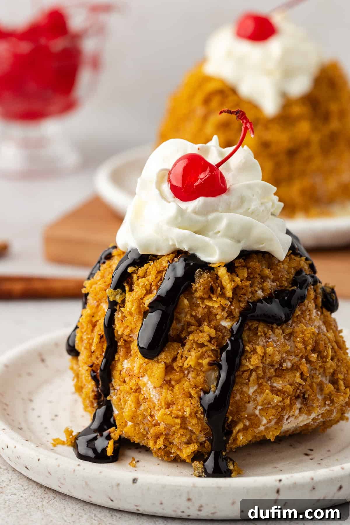 A beautifully presented fried ice cream ball, generously drizzled with rich chocolate syrup, crowned with a cloud of whipped cream, and finished with a bright maraschino cherry. Another plate of fried ice cream and a bowl of cherries are softly visible in the background.