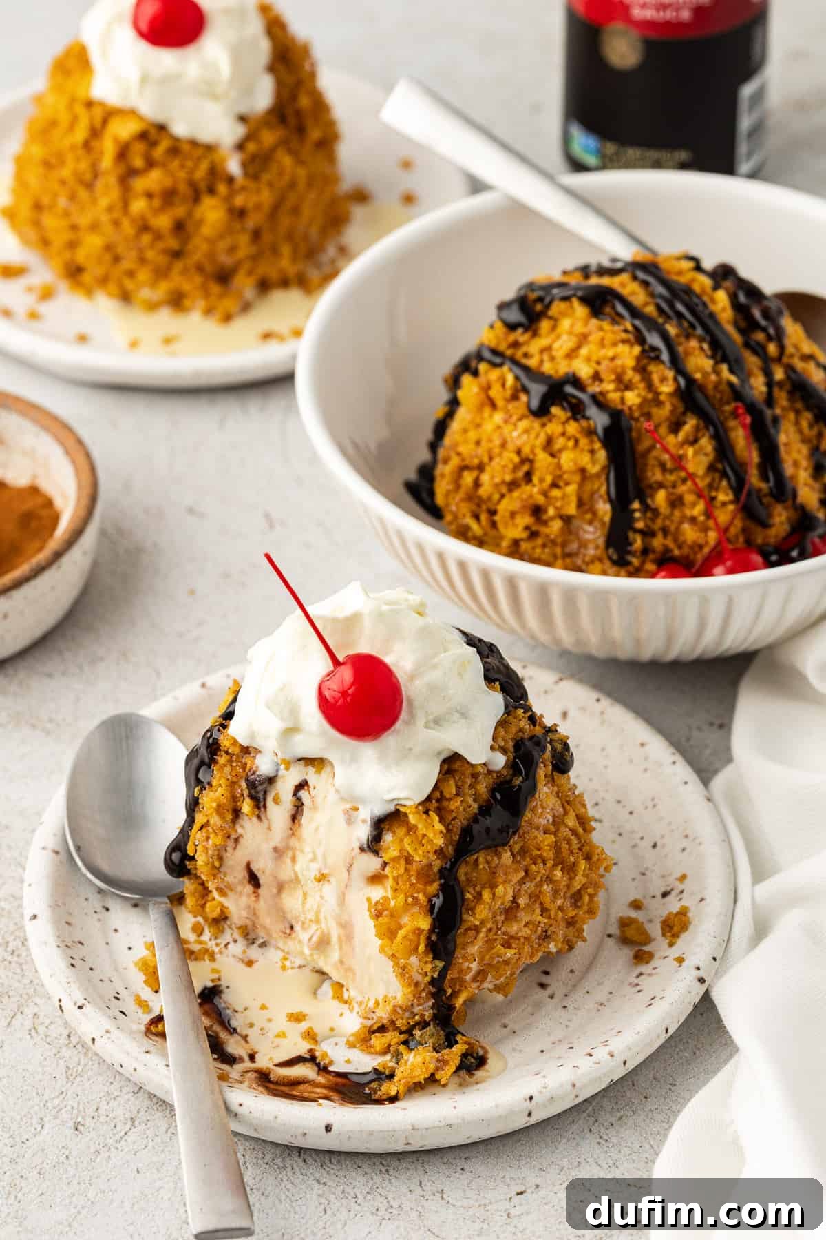 A partially eaten ball of fried ice cream on a small white plate with a spoon, accompanied by a bowl holding another ice cream ball, a crisp white towel, and another ice cream portion in the background, creating a cozy and inviting dessert scene.