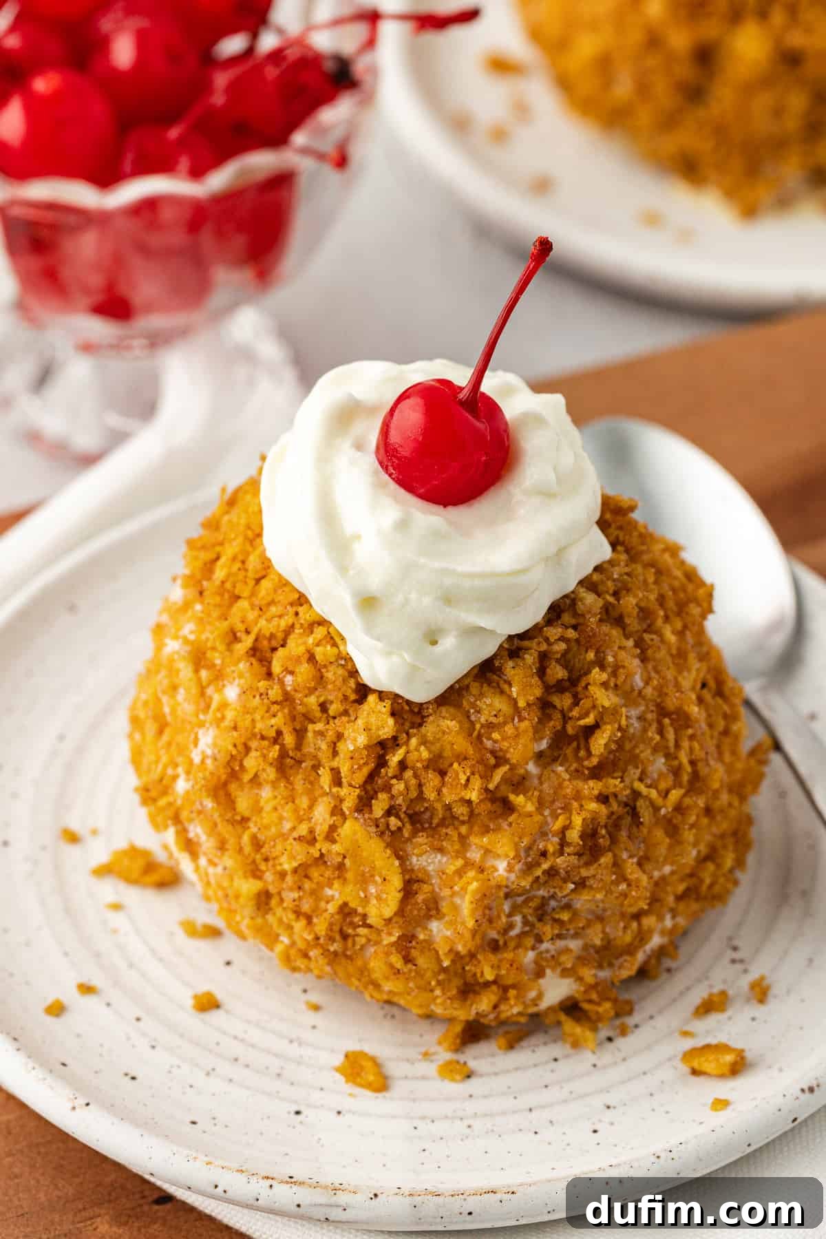 A single ball of perfectly 'fried' ice cream, artfully topped with whipped cream and a cherry, sits on a small white plate with a spoon. A bowl of additional cherries and more fried ice cream balls can be seen in the soft background, inviting a delightful dessert experience.