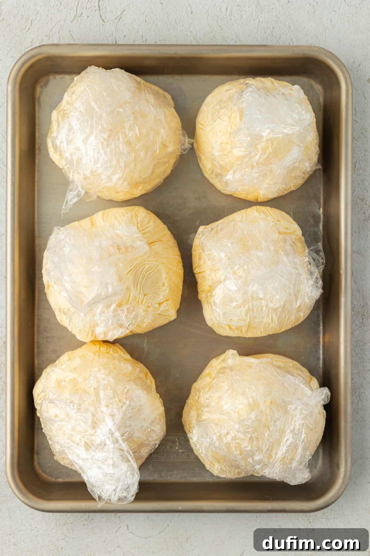 Six perfectly formed vanilla ice cream balls, individually wrapped in plastic, neatly arranged in two rows of three on a metal baking sheet, ready for the freezer.