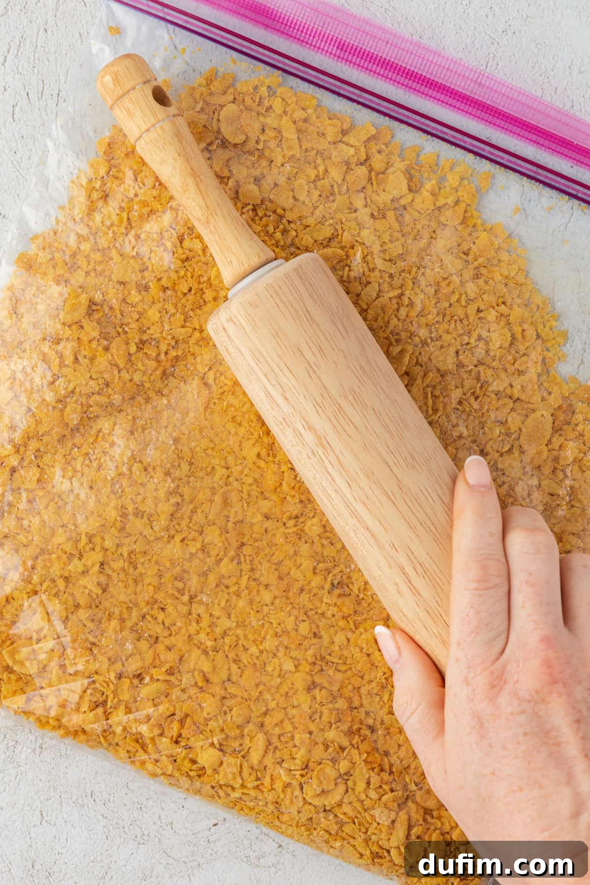 A hand is actively using a wooden rolling pin to crush cornflakes inside a clear ziploc bag, preparing them for the crispy coating.