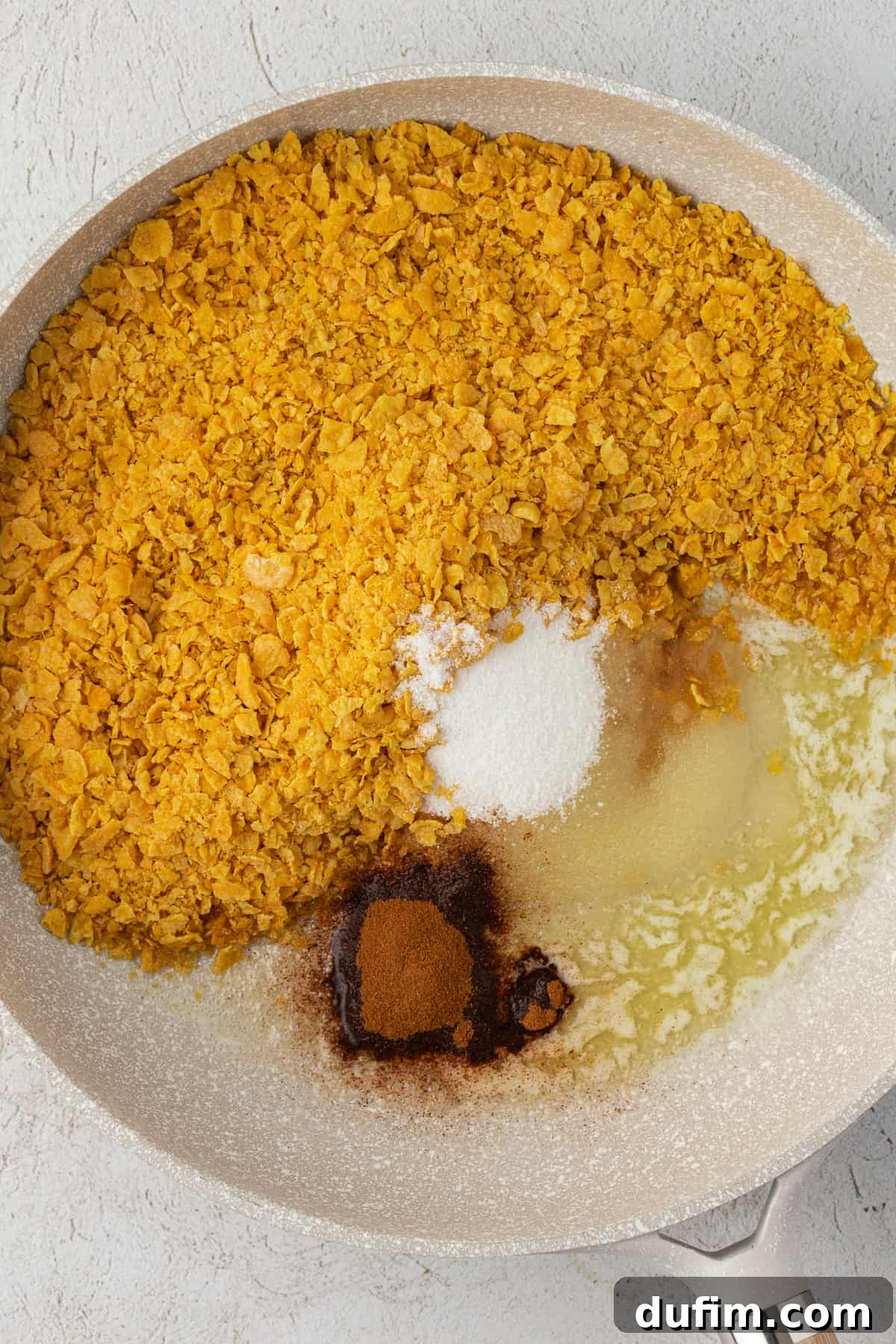 A medium skillet filled with crushed corn flakes, melted butter, granulated sugar, cinnamon, and vanilla extract, ready to be combined into the aromatic coating mixture.