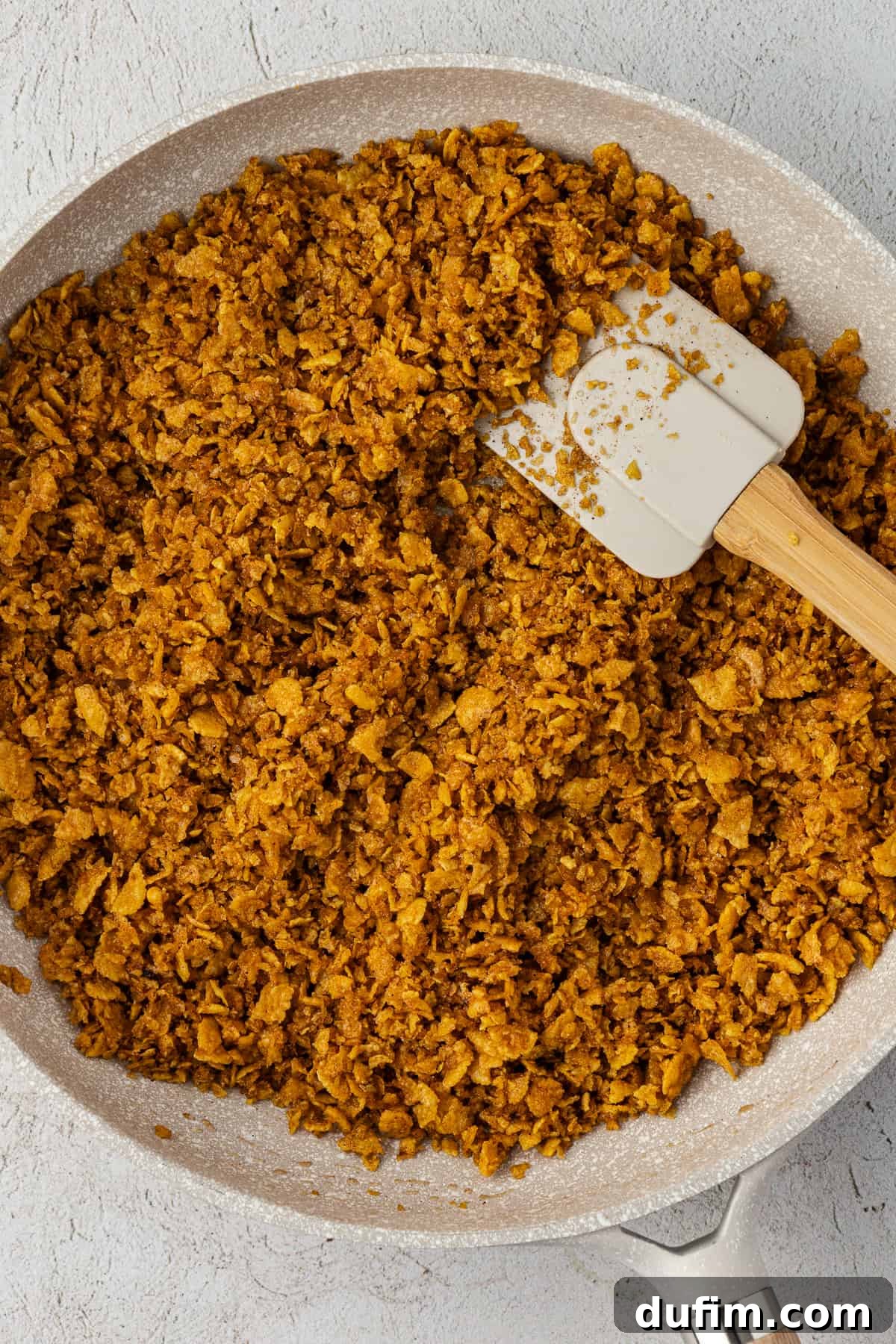 A spatula stirs the rich, toasted cornflake cereal mixture in a pan, highlighting its appealing golden-brown color.