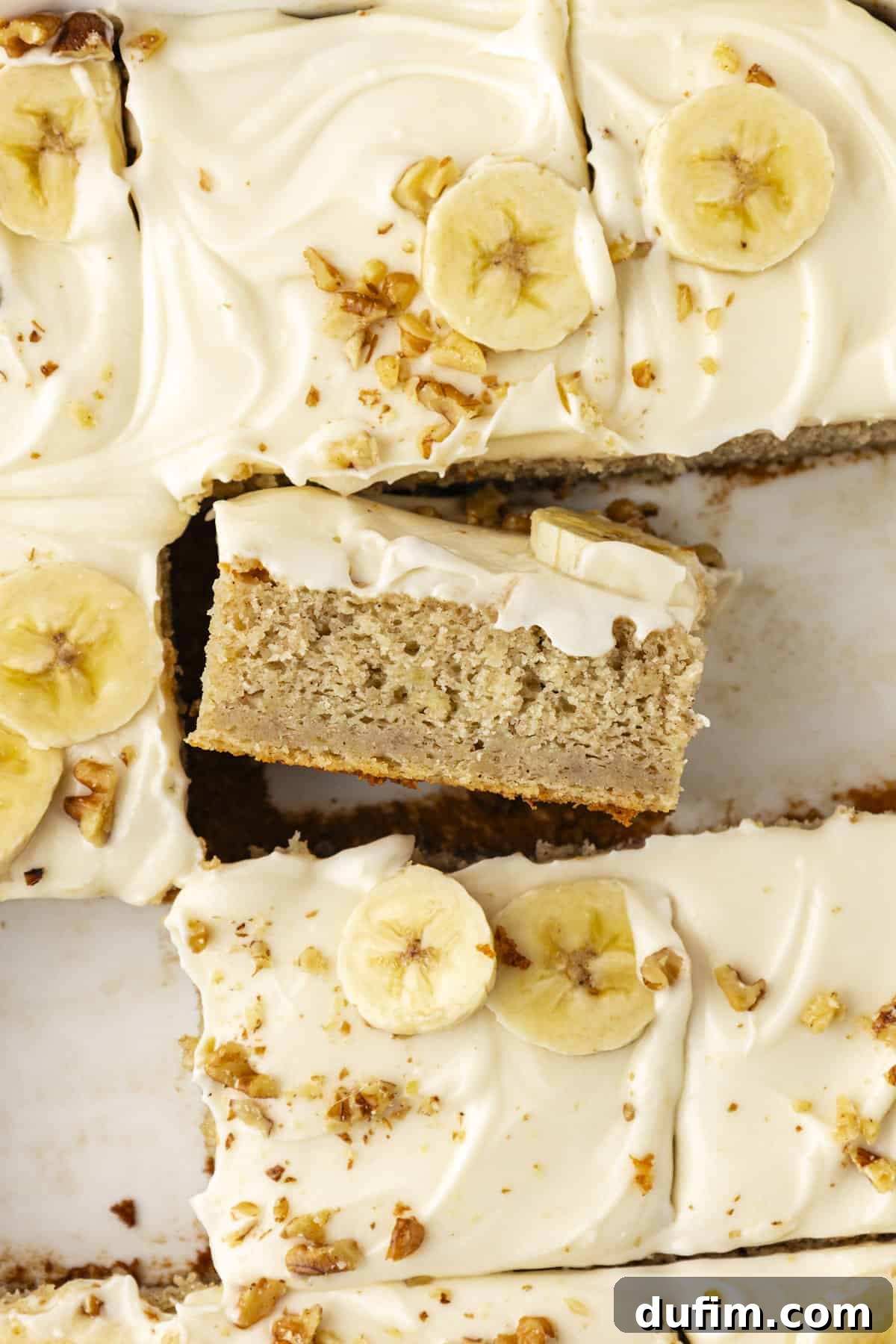 Close up of a moist banana cake with some slices missing and one slice turned on its side to reveal the layers of cake and generous cream cheese frosting
