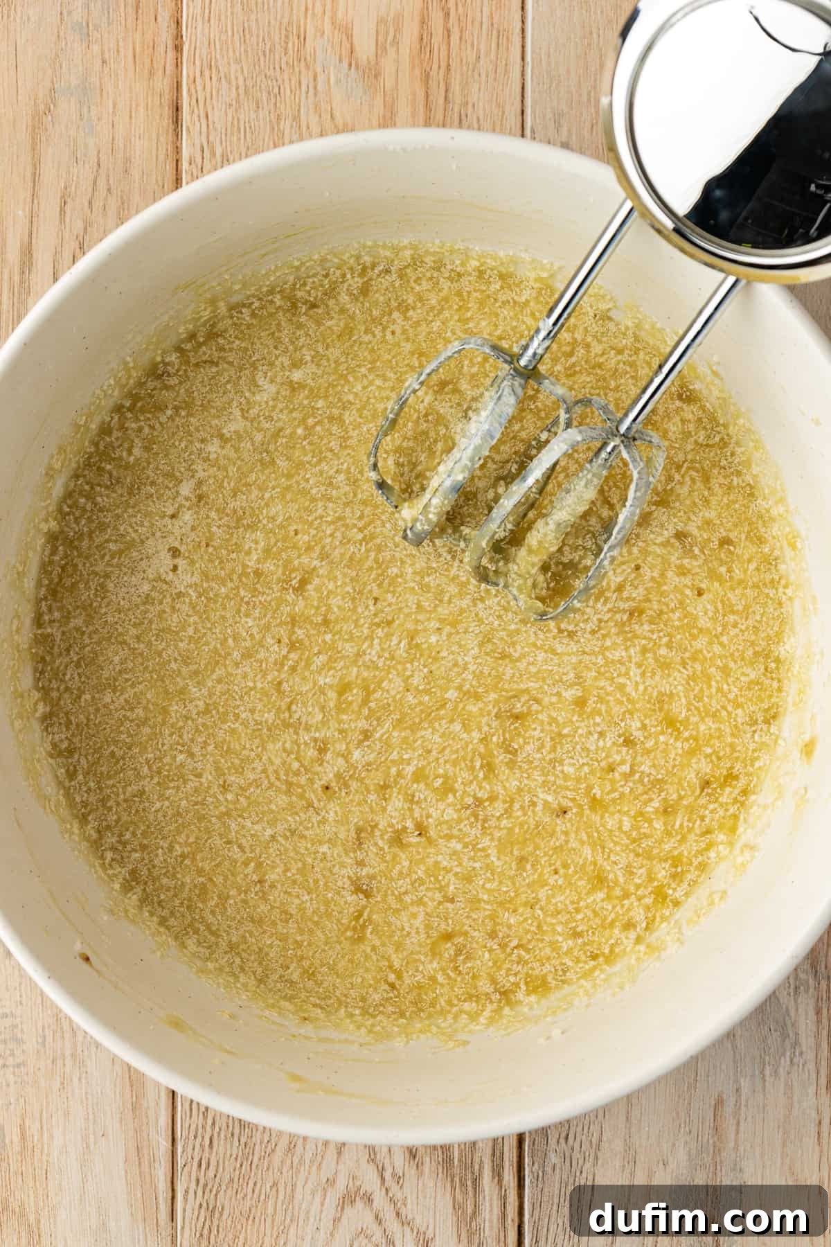 A white bowl on a wood surface containing a fully mixed banana cake wet ingredient mixture, with an electric mixer resting inside the bowl